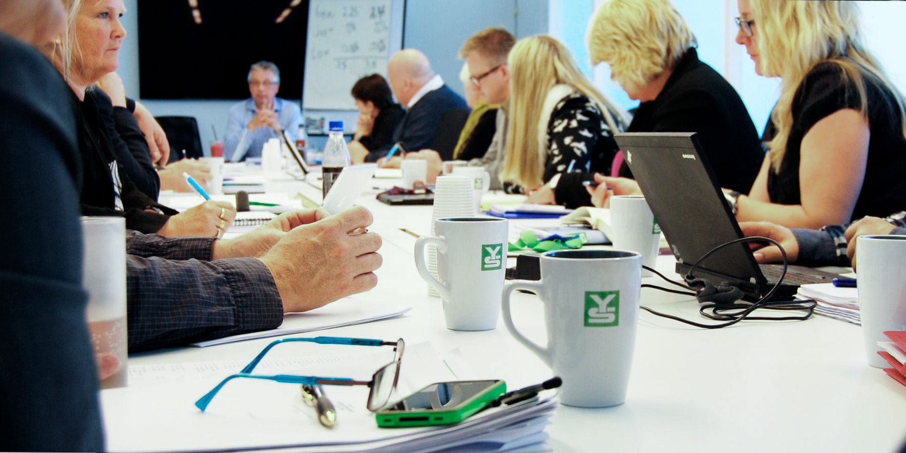 Kopper med YS-logo. Folk sitter rundt et møtebord.