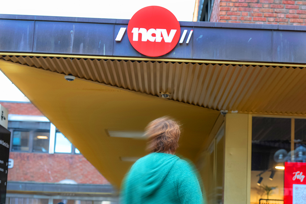 Person i grønn genser står foran bygning med rør nav-logo
